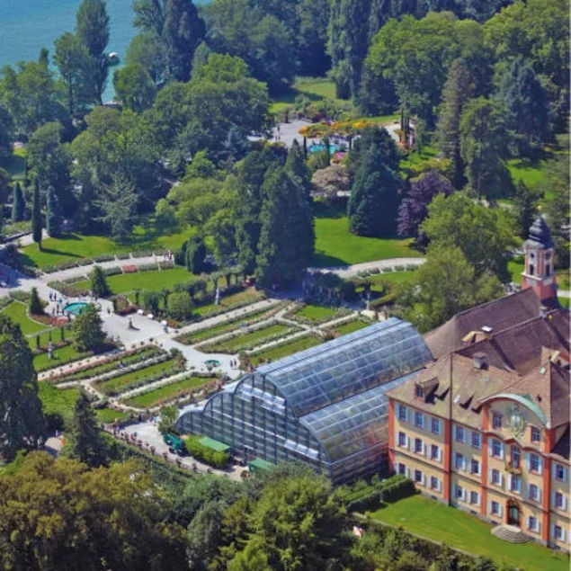 Luftaufnahme einer weitläufigen Parkanlage mit gepflegten Gartenflächen, einem großen Gewächshaus aus Glas und einem historischen Gebäude mit rotem Dach, umgeben von vielen Bäumen.