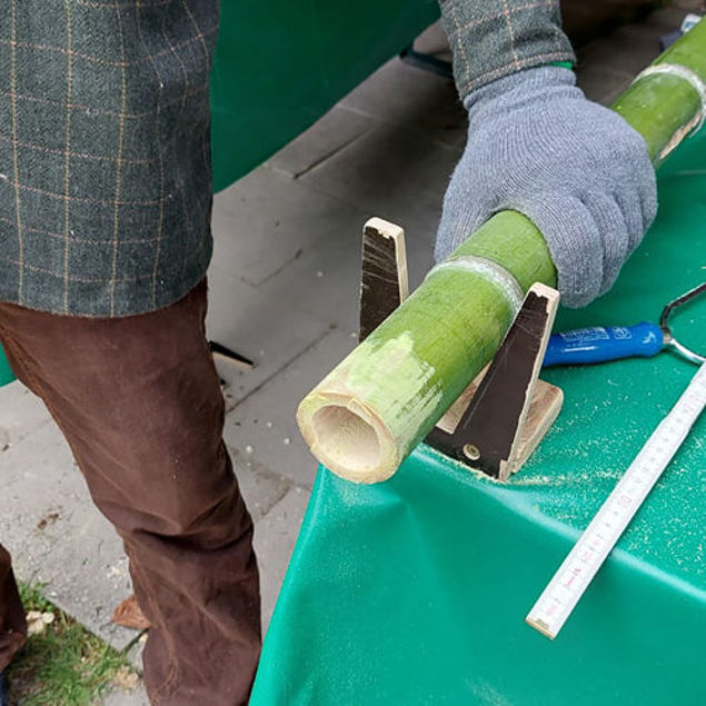 Person mit Handschuhen sägt ein grünes Bambusrohr auf einem Arbeitstisch, daneben liegen Werkzeuge wie Säge und Zollstock
