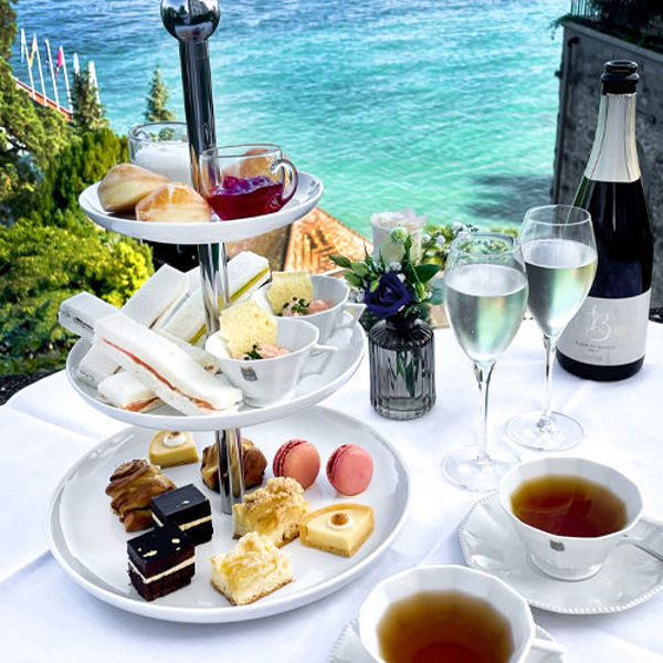 Auf einem Tisch mit weißer Tischdecke steht eine Etagere. Darauf befinden sich verschiedene Kuchen. Außerdem stehen 2 Tassen Tee, zwei Sektgläser und eine Sektflasche auf dem Tisch. Im Hintergrund ist Wasser zu sehen.