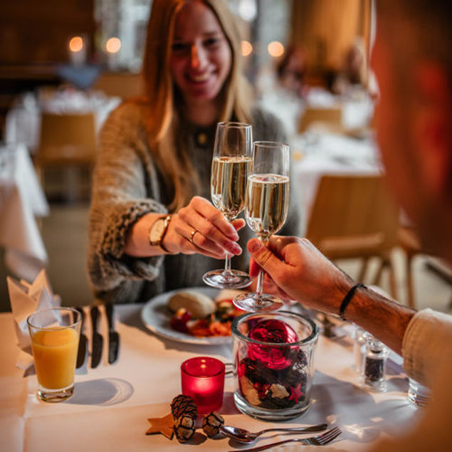 Zwei Personen stoßen mit Sektgläsern an, während sie an einem festlich gedeckten Tisch sitzen. Auf dem Tisch sind ein Teller mit Essen, ein Glas Orangensaft und eine dekorative Glasvase mit roten und schwarzen Elementen zu sehen.