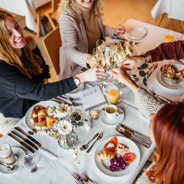 Eine Gruppe von Menschen sitzt an einem gedeckten Tisch. Die Personen lachen sich an und stoßen mit einem Glas Prosecco an. Auf dem Tisch gibt es Latte Macchiato, Kaffee, Orangensaft; auf den Tellern liegen Croissants, Obst und Müsli